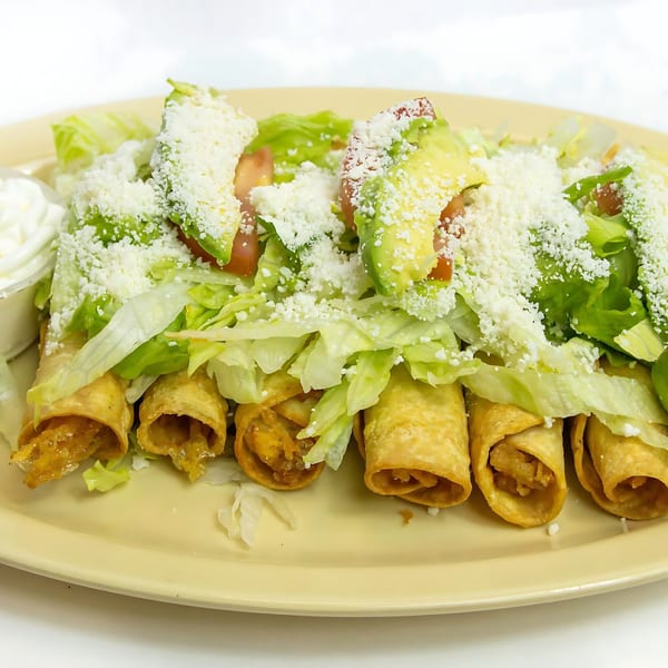 Five golden-brown taquitos topped with shredded lettuce, diced tomatoes, avocado slices, and crumbled cheese.
