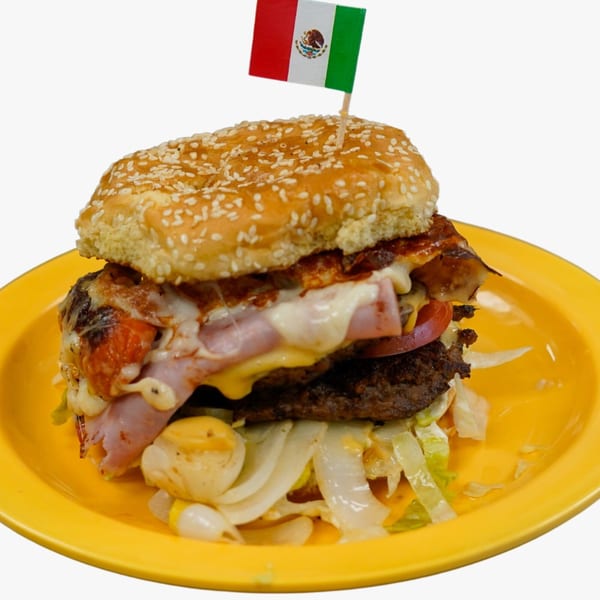 A large burger with cheese, ham, bacon, and vegetables, topped with a Mexican flag pick.