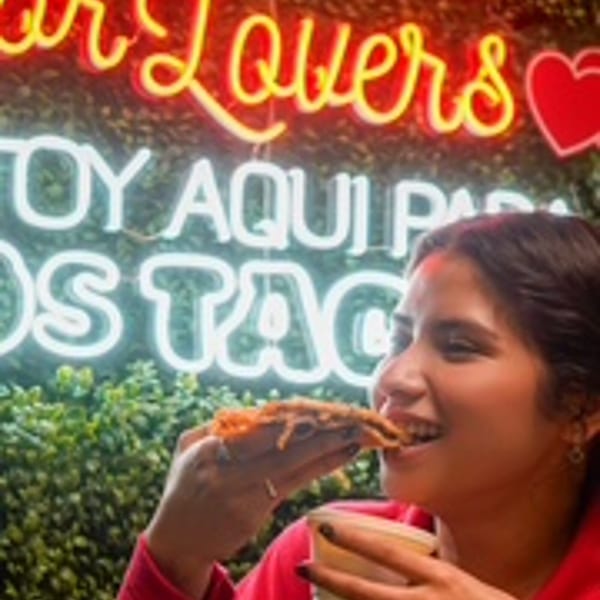 Woman happily eating a large taco in front of neon signs at Taco Riendo Taqueria.