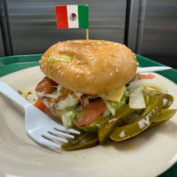 A Mexican torta with a flag on top, served with pickled jalapeños and a white plastic fork.