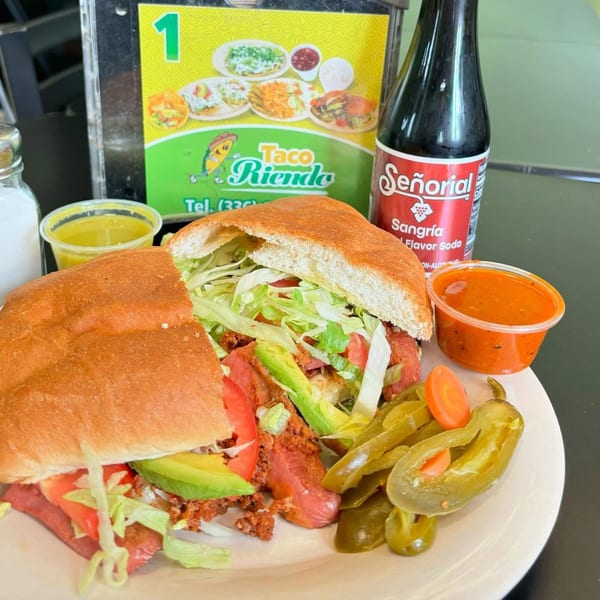 A large Mexican torta sandwich with avocado, lettuce, and tomato, served with pickled peppers and a soda.