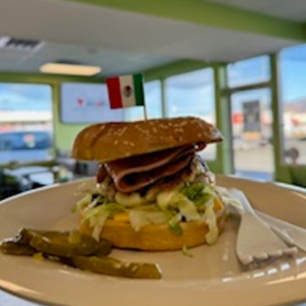 A Mexican torta with a flag toothpick, lettuce, cheese, and pickles on a white plate.