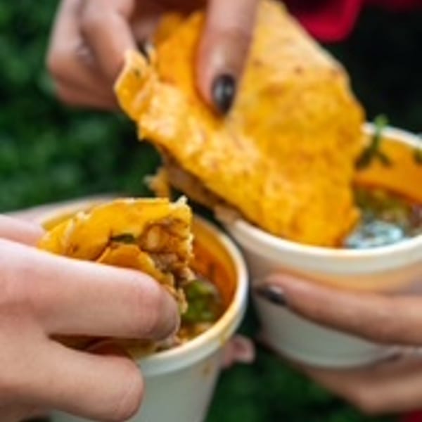 Two hands holding birria tacos with consommé in white cups, ready to eat.