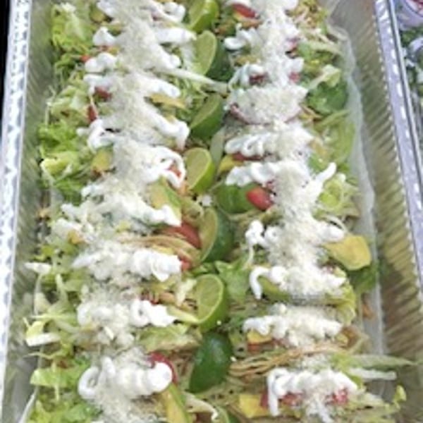 A catering tray filled with many tacos topped with lettuce, cheese, sour cream, avocado, and lime.