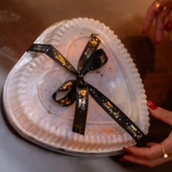 Heart-shaped dessert in a clear container with a black and gold ribbon, held by hands.