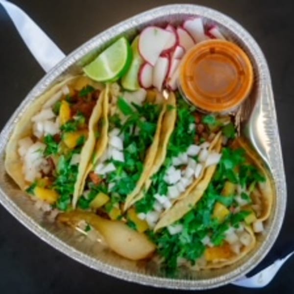 Three street tacos with cilantro, onions, lime, radishes, and sauce in a foil container.