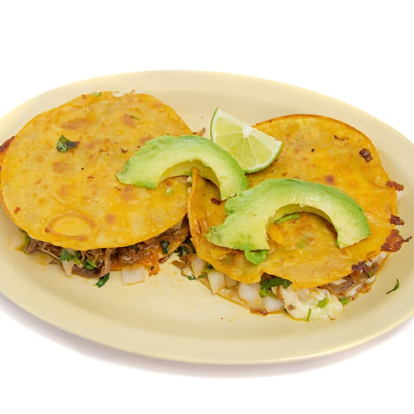 Two birria tacos with melted cheese, shredded meat, onions, cilantro, avocado slices, and a lime wedge.