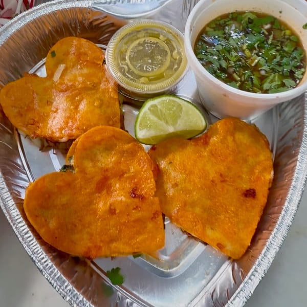 Three heart-shaped birria tacos with consommé, lime, and salsa in a foil container.