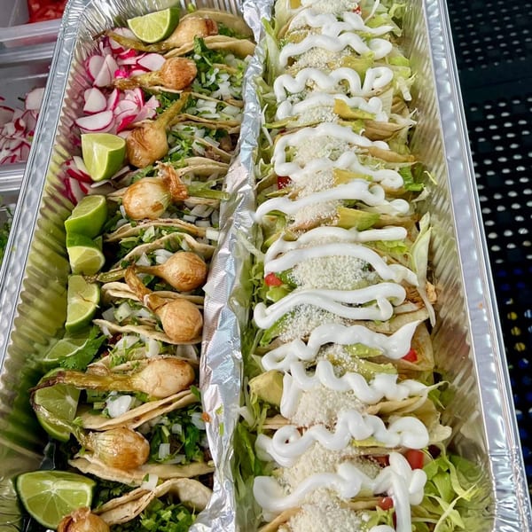 Two foil trays filled with various tacos, one with radishes and limes, the other with sour cream and cheese.