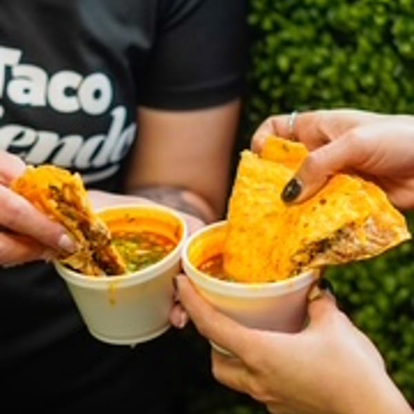 Two hands dipping birria tacos into cups of consommé, with a person wearing a "Taco Riendo" shirt.