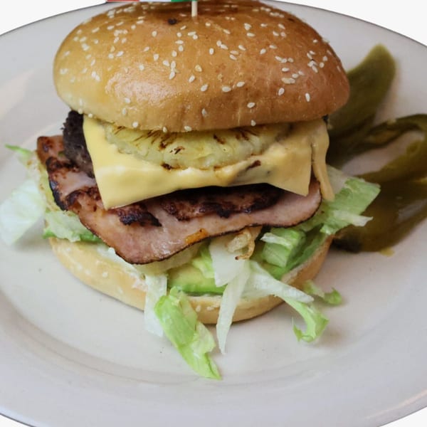 A hamburger with pineapple, cheese, ham, and lettuce on a sesame seed bun, served with pickles.