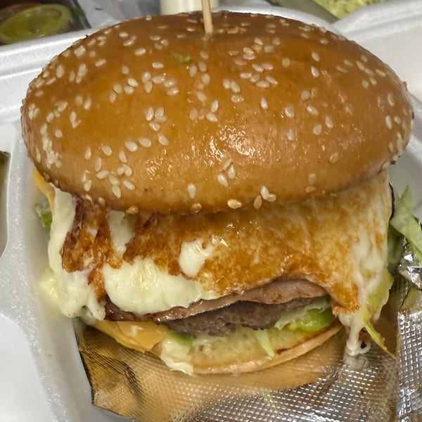 A close-up of a burger with melted cheese, lettuce, and a sesame seed bun in a takeout container.