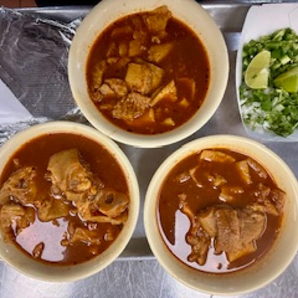Three bowls of rich, red menudo soup with meat, alongside a plate of chopped onions and lime.