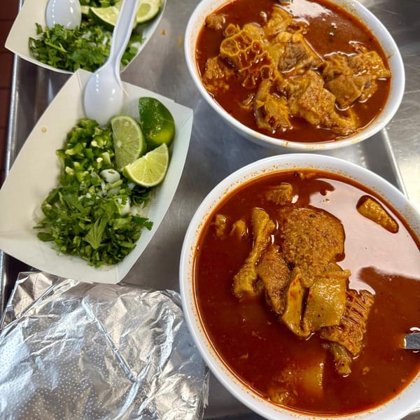 Two bowls of menudo soup with tripe, served with sides of cilantro, onions, jalapeños, and lime.