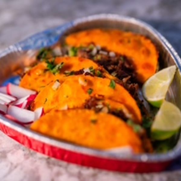 Four birria tacos with cilantro, onions, lime wedges, and sliced radishes in a foil container.