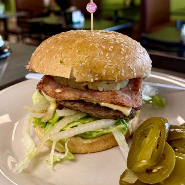 A burger with bacon, cheese, lettuce, and onions, served with pickled jalapeños on a white plate.