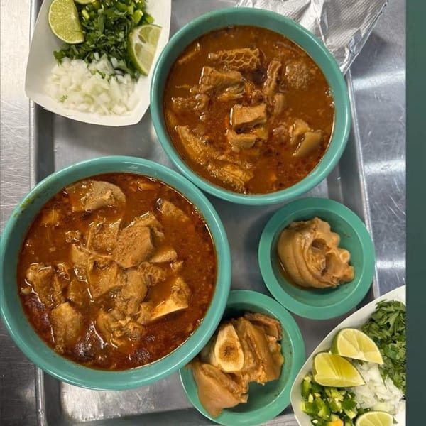 Two bowls of menudo soup with sides of chopped onions, cilantro, jalapeños, and lime wedges.